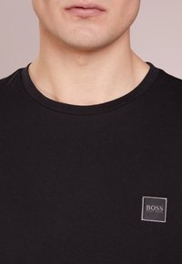 Camiseta de algodón negra con cuello redondo y un pequeño logo bordado cuadrado en gris en el lado izquierdo del pecho. Textura suave.