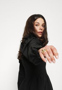 Rochie neagră cu un corset plisat, mâneci ondulate și fustă evazată. Inel de aur pe mână, piele deschisă și păr ondulat de culoare brown.