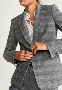 Veste à carreaux grise en tissu texturé, avec une fermeture à deux boutons, des revers crantés et des lignes contrastantes bleu et marron.