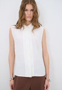 Mouwloze witte blouse van lichtgewicht stof met een voorknoopsluiting en subtiele glans, gecombineerd met een paarse broek en een eenvoudige armband.