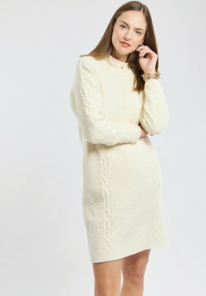 Robe pull en tricot torsadé crème, présentant des motifs texturés, des manches longues et un col rond. Longueur genoux avec coupe ample.