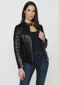 Veste en cuir noir au design ajusté, avec des panneaux matelassés horizontaux, un col court et une fermeture éclair. Associée à un jean en denim foncé.