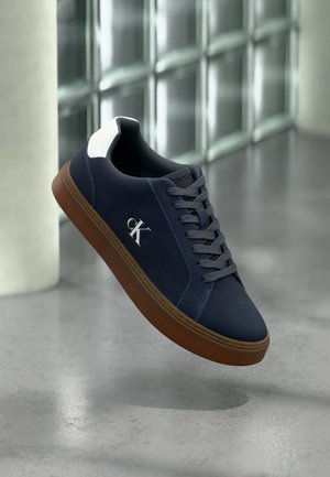 Sötétkék sportcipő barna talppal, szürke fűzőkkel, fehér sarokdíszítéssel és fehér "CK" logóval, beltéren lebegve üvegblokkokkal tarkított fal és betonpadló közelében.