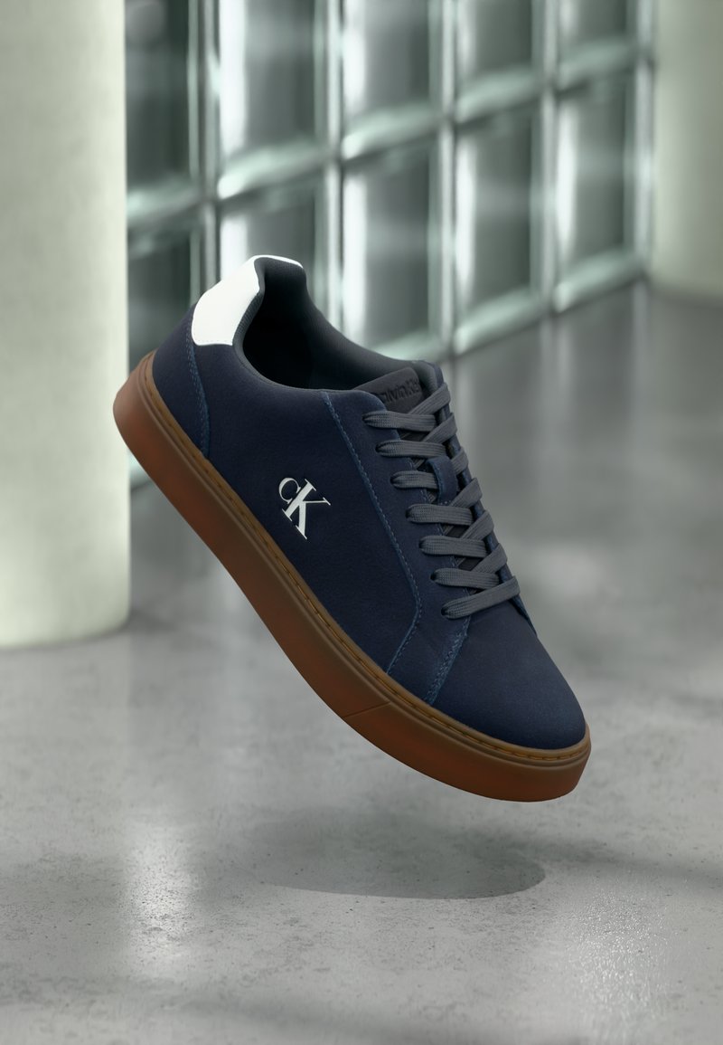 Μπλε ναυτικό αθλητικό παπούτσι με καφέ σόλα, γκρι κορδόνια, λευκή λεπτομέρεια στη φτέρνα και λευκό λογότυπο "CK", αιωρείται σε εσωτερικό χώρο κοντά σε τοίχο από γυάλινα τουβλάκια και σκυρόδεμα στο πάτωμα.