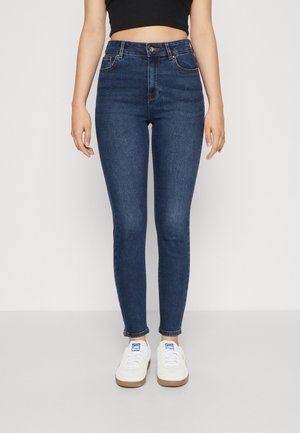 Slanke mørkeblå jeans båret af en person, der står, kombineret med hvide sneakers og en sort cropped top.