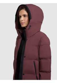 Jachetă căptușită burgundy cu guler înalt și glugă mare. Are accente negre, buzunare cu fermoar și o textură netedă, lucioasă.