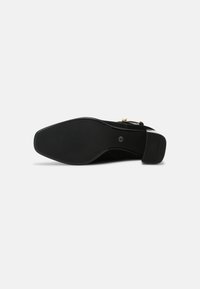 Semelle noire d'une chaussure à talon pour femme avec un bout carré et un talon texturé, sur un fond gris clair uni.