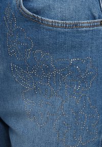 Μπλε τζιν παντελόνι με floral σχέδιο δημιουργημένο από ασημένιες στρας στην περιοχή της τσέπης, με υφή ομαλή φινίρισμα.