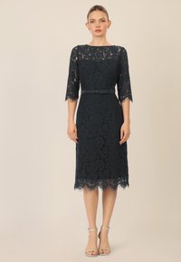 Vestido de encaje en un azul marino oscuro con mangas de tres cuartos, dobladillo con picos y una cintura ajustada acentuada por un cinturón a juego. Tacones plateados con tiras.