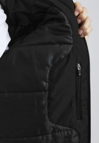Interiorul jachetei puf negre, cu un material neted și lucios, dotat cu un buzunar cu fermoar și o textură subtilă quilted.