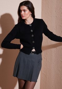 Black cardigan with button closure and textured finish, paired with a pleated gray skirt. Model stands against a light-colored wall.