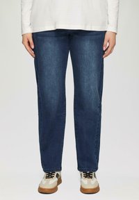 Donkerblauwe denim jeans met een recht model. Het model draagt beige en grijze sneakers met zwarte veters.