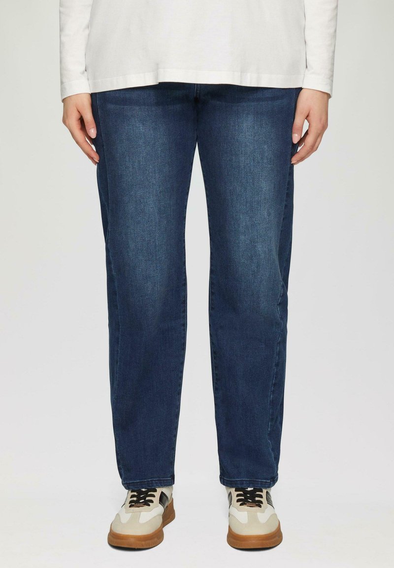 Donkerblauwe denim jeans met een recht model. Het model draagt beige en grijze sneakers met zwarte veters.