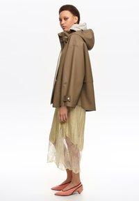 Person wearing a brown hooded rain jacket, yellow lace midi skirt, and pink low-heeled shoes, standing in profile against white background.