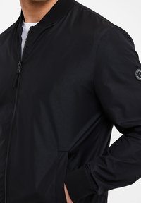 Chaqueta bomber negra con una textura suave y brillante. Cuenta con un cierre de cremallera en la parte frontal, cuello y puños acanalados, y un parche en la manga izquierda.
