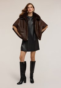 Žena v černých kožených mini šatech se zdobením, tmavě hnědém beránkovém kabátu a černých kozačkách na podpatku, stojící s rukama na bocích.