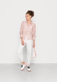 Camisa rosa de renda com hem lisado, combinada com calças brancas e ténis em tons pastel. Segura uma pequena bolsa rosa claro.