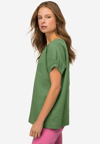 Top in lino verde con maniche corte arricciate e una vestibilità comoda. Presenta un ampio scollo, abbinato a pantaloni rosa.