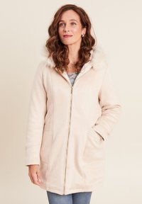 Beige gewatteerde jas met een faux fur rand, ritsluiting, zijzakken en een zachte textuur, gedragen met een lichtgrijze trui en blauwe jeans.