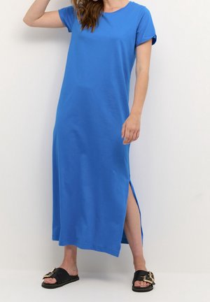 Frau trägt ein blaues Maxikleid mit kurzen Ärmeln und Seitenschlitz sowie schwarze Slide-Sandalen mit goldenen Schnallen, stehend vor einem einfarbigen weißen Hintergrund.