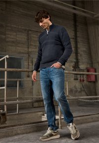 Tmavě modrý svetr na zip s texturovanými pruhy, kombinovaný s světle modrými džínami a šedými teniskami. Má stojáček a žebrované manžety.