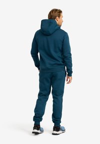 Teal hoodie och matchande joggers, tillverkade av mjukt tyg. Har en huva, elastiska ärmslut och sidofickor, bärs med svarta och blå sneakers.