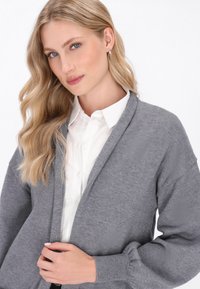 Hellgrauer Cardigan mit langen Ärmeln, offener Vorderseite und gerippten Bündchen. Über einem weißen Button-Up-Hemd getragen, das eine glatte Textur und ein schlichtes Design aufweist.