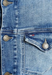 Jeansjacka i ljusblått tyg med en sliten textur, prydd med silverknappar och en liten röd, vit och blå flik.