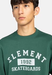 Zelená mikina s nápisem "ELEMENT", "1992" a "SKATEBOARDS" v bílé barvě. Hladký límec a strukturovaná tkanina. Uvolněný střih.