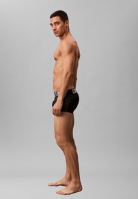 Zwarte boxershorts met een logo-elastiek, met een nauwsluitende pasvorm, gemaakt van rekbaar materiaal. Het onderwerp staat zijwaarts geposeerd, op blote voeten.