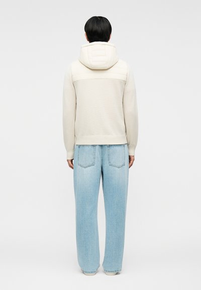 Άτομο που φαίνεται από πίσω φορώντας μια κρεμ φούτερ με κουκούλα και επένδυση στο πάνω μέρος της πλάτης, καθώς και ανοιχτομπλε τζιν παντελόνι με φαρδιά πόδια.