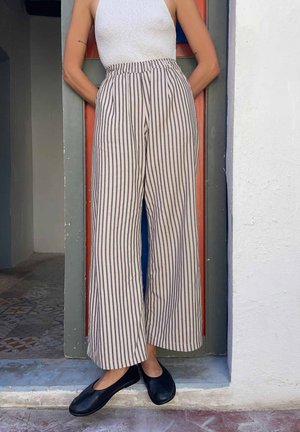 Persoon draagt hoge taille, wijde gestreepte broek, wit mouwloos topje en zwarte platte schoenen, staand in een deurpost met handen in de zakken.