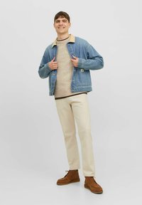 Veste en jean avec un col beige, pull crème, pantalon clair et chaussures marron. Le mannequin se tient avec les mains sur la veste, souriant.
