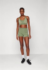 Sportovní podprsenka olivově zelená a odpovídající krátké šortky s vysokým pasem z elastické tkaniny. Navrženo pro pohodlí a volnost pohybu. Nohy obuty v černých sportovních botách.