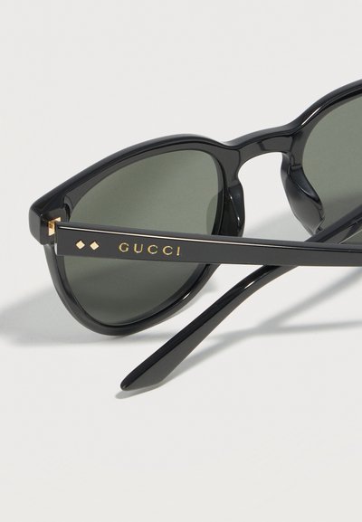 Μαύρα γυαλιά ηλίου με σχήμα γατίσιου ματιού, σκούρους φακούς και χρυσό λογότυπο Gucci στο πλαϊνό μπράτσο, τοποθετημένα σε λευκή επιφάνεια.