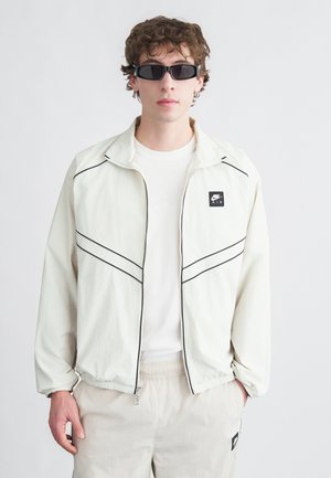 Giacca leggera color crema con dettagli neri, chiusura zip e logo Nike. Indossata sopra una t-shirt bianca, abbinata a pantaloncini elasticizzati beige.