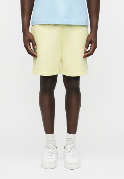 Man wearing pale yellow shorts, light blue shirt, white socks, and white sneakers, standing against a plain white background.