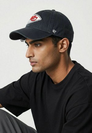 Man wearing black Kansas City Chiefs baseball cap and black t-shirt, looking to the left against a plain background.