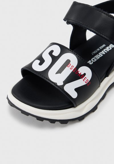 Μαύρα σανδάλια-σχέδιο slide από συνθετικό υλικό, με φαρδιά λουρί που φέρει λευκό κείμενο "SQ2 DSQUARED2" και υφασμένη καουτσούκ σόλα.