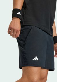 Čierne športové šortky vyrobené z texturovanej tkaniny, so bočnými vreckami a bielym logom Adidas s tromi pruhmi na dolnej ľavej strane.