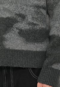 Κοντινό πλάνο ενός ατόμου που φοράει έναν γκρι υφασμάτινο πουλόβερ με ραμμένα μανίκια και στρίφωμα, συνδυασμένο με μαύρο παντελόνι που έχει λευκές ραφές.