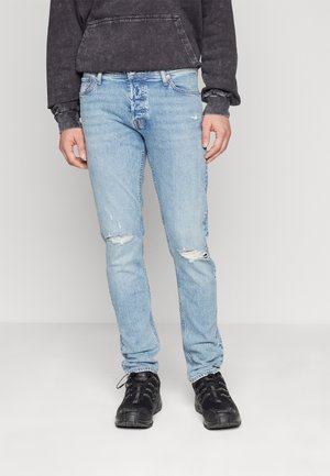 Muž oblečený v světle modrých roztrhaných džínách, černých teniskách a tmavě šedé vyprané mikině s kapucí, stojící na jednoduchém světlém pozadí.