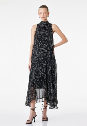 Femme debout portant une robe midi noire sans manches à pois blancs et des sandales à talons hauts noires à brides, sur fond blanc.