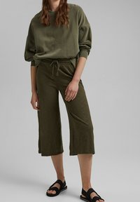 Felpa oversized di colore verde oliva con scollatura a coste e spalle scese, abbinata a pantaloni corti con coulisse in coordinato e sandali flatti.