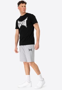 Čierne tričko s krátkym rukávom s bielym logom "TAPOUT" v kombinácii so sivými šortkami s rovnakým logom. Na sebe má biele tenisky a ponožky.