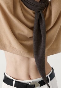 Pull court beige noué à la poitrine avec une écharpe marron foncé, dévoilant le ventre, associé à un pantalon blanc et une ceinture noire avec une boucle argentée.