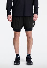 Pantalones cortos de correr negros con forro de compresión incorporado, tela liviana y un logo reflectante. Llevados con zapatos deportivos negros y calcetas.