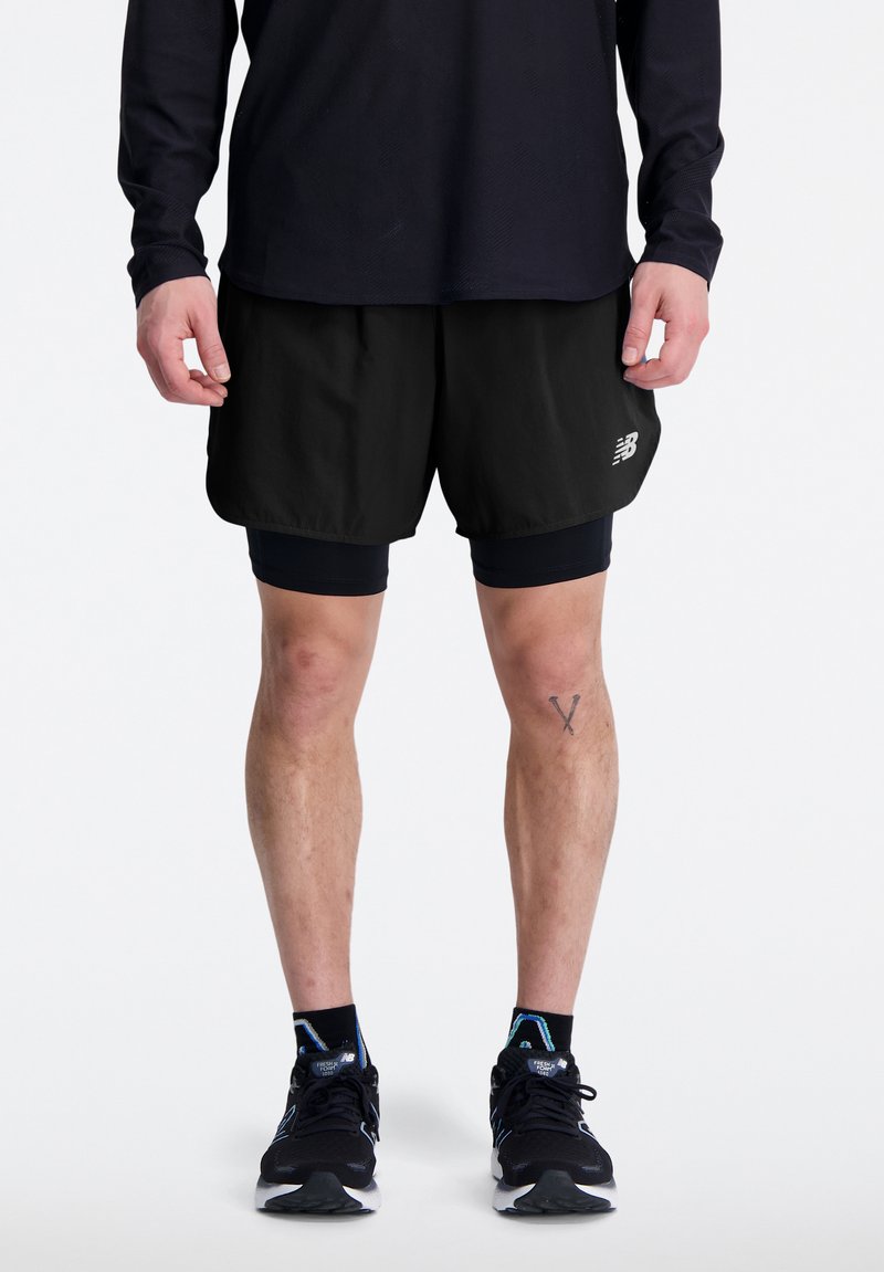 Pantalones cortos de correr negros con forro de compresión incorporado, tela liviana y un logo reflectante. Llevados con zapatos deportivos negros y calcetas.