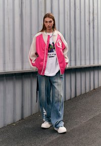 Chaqueta oversized rosa y crema, camiseta gráfica blanca, jeans holgados azul desgastado y zapatillas blancas sin cordones. Detalle de collar de cadena visible.