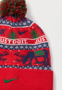 Κόκκινο πλεκτό σκουφάκι με πράσινη πομ-πομ. Διακοσμημένο με λευκά και ναυτικά σχέδια, δέντρα και σχέδιο με ελάφι. Η φράση "JUST DO IT" σε λευκό κείμενο και πράσινο λογότυπο Nike.